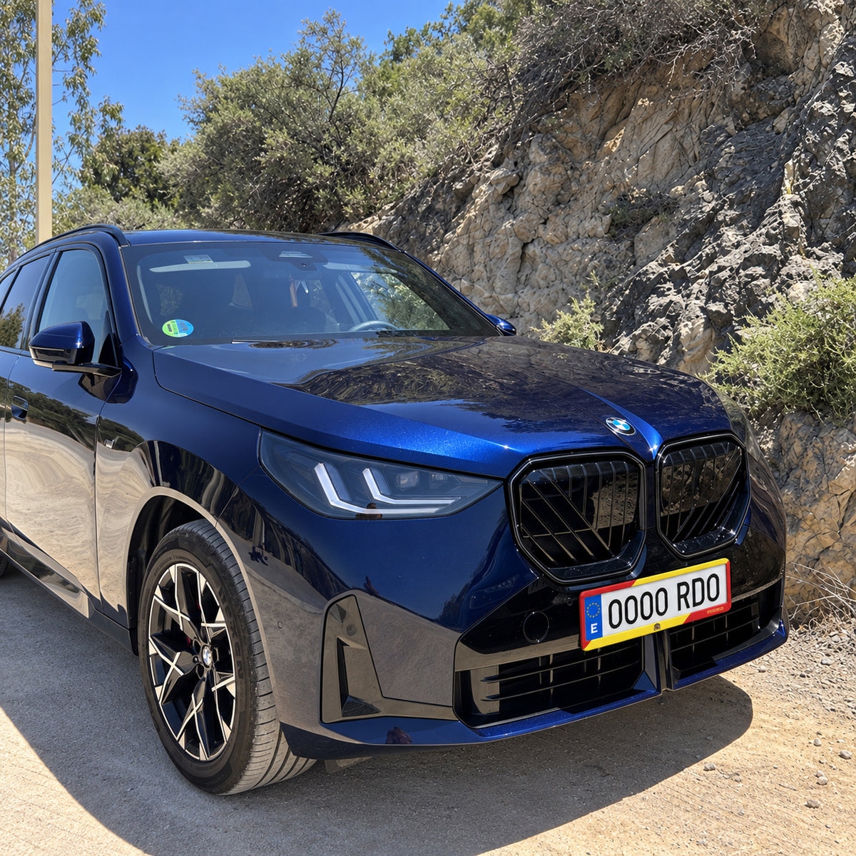 Portamatrículas Rodeo España Race instalado en un BMW azul junto a una carretera rocosa.